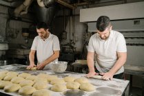 Зрелые бородатые этнические пекари формируют хлеб из теста за столом с мукой и миской в пекарне — стоковое фото
