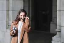 Стильна жінка в пальто і сонцезахисних окулярах стоїть з старовинною фотокамерою на вулиці і дивиться на камеру — стокове фото