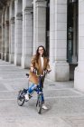 Mujer alegre en abrigo mirando hacia otro lado en bicicleta contra el viejo edificio con columnas en la ciudad - foto de stock