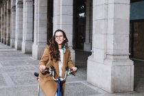 Femme gaie en manteau regardant loin sur le vélo contre vieux bâtiment avec des colonnes en ville — Photo de stock