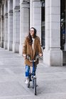 Femme gaie en manteau regardant loin sur le vélo contre vieux bâtiment avec des colonnes en ville — Photo de stock
