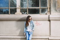Молодая женщина в модном джинсовом наряде и солнцезащитных очках, стоящая с рукой в кармане возле потрепанного здания в городе — стоковое фото