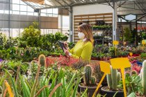 Busy female gardener in mask standing near various plants and browsing tablet while working in garden center — Stock Photo