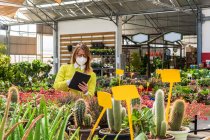 Занятая женщина-садовница в маске стоит рядом с различными растениями и просматривает планшет во время работы в центре сада — стоковое фото