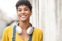 Mujer afroamericana sonriente soñadora con auriculares inalámbricos mientras está de pie en la calle mirando la cámara - foto de stock