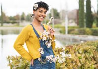 Gioiosa femmina afroamericana in piedi con rami con fiori su tutto il corpo in parco in primavera e guardando la fotocamera — Foto stock