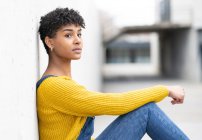 Vista lateral de una mujer afroamericana positiva en overol denim y con peinado afro apoyado en la pared y mirando hacia otro lado - foto de stock