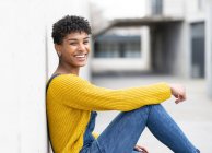 Side view of positive African American female in denim overalls and with Afro hairstyle leaning on wall and looking at camera while sitting on city street — Stock Photo