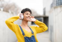 Dreamy smiling African American female in wireless headphones enjoying songs with closed eyes while standing on street — Stock Photo