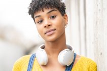 Mujer afroamericana sonriente soñadora con auriculares inalámbricos mientras está de pie en la calle mirando la cámara - foto de stock