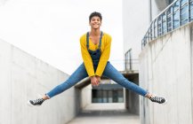 Vue latérale d'une femme afro-américaine active et joyeuse portant des salopettes et des baskets au moment de sauter au-dessus de la rue de la ville — Photo de stock