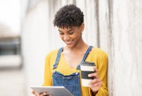 Frohe schwarze Frau, die mit Coffee to go in Pappbecher auf der Straße steht und Videos auf dem Tablet anschaut, während sie am Wochenende in der Stadt unterhält — Stockfoto