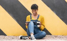 Прохладный афроамериканка сидит с напитком на вынос в бумажной чашке возле стены в городе и серфинг Интернет на планшете — стоковое фото