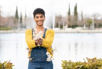 Радостная афроамериканка, стоящая с ветками с цветами в парке весной и смотрящая в камеру — стоковое фото