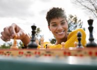 From below of optimistic African American female sitting at table in park and playing chess while looking at camera — Stock Photo