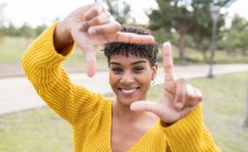 Mulher afro-americana positiva com cabelo encaracolado em pé no parque e mostrando sinal de enquadramento enquanto olha para a câmera — Fotografia de Stock