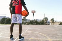 Vista trasera recortada de un irreconocible jugador de streetball afroamericano en uniforme de pie con pelota en la cancha de baloncesto - foto de stock