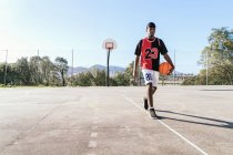 Seriöser afroamerikanischer Streetballspieler in Uniform läuft mit Ball auf Basketballfeld und schaut in die Kamera — Stockfoto
