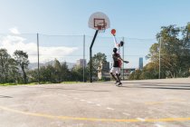 Vista lateral do amigo afro-americano masculino e hispânico jogando streetball enquanto salta e marca bola no aro — Fotografia de Stock