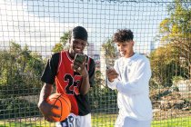 Sonrientes jugadores de streetball afroamericanos de pie cerca de la red en el patio de baloncesto y navegando por teléfonos móviles - foto de stock