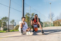 Sonrientes jugadores de streetball afroamericanos sentados en el patio de baloncesto en un día soleado y mirándose unos a otros - foto de stock