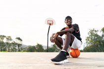 Low angle of smiling black male streetball player sitting on ball on basketball playground and looking away — Stock Photo