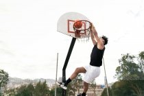 Vista lateral del jugador de streetball masculino étnico realizando slam dunk en el momento de saltar por encima del patio de recreo y anotar baloncesto en el aro - foto de stock