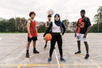 Confident multiethnic streetball team with ball standing on basketball sports ground in city looking at cmaera — Stock Photo