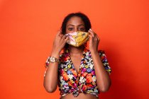 Привлекательная молодая афроамериканка в красочном топе надевает тканевую маску для лица и смотрит на камеру на красном фоне — стоковое фото