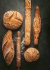 Top view composition with various typed of freshly baked crusty artisan bread loaves of different shapes placed near wooden rolling pin on black background — Stock Photo