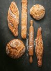 Composición de vista superior con varios tipos de panes artesanales recién horneados de diferentes formas colocados cerca de un rodillo de madera sobre fondo negro - foto de stock