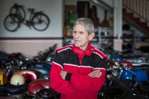Серйозний старший чоловічий механік у червоному робочому одязі, що стоїть у майстерні ремонту проти пошкоджених іржавих мотоциклів — стокове фото
