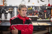 Серьезный старший механик в красной рабочей одежде, стоящий в ремонтной мастерской с профессиональными инструментами и инструментами, отводящими взгляд — стоковое фото