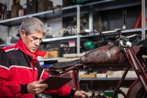 Schwere grauhaarige ältere Reparaturarbeiter mit Tablet und Videoanleitung beim Reparieren alter kaputter Motorräder in der Garage — Stockfoto