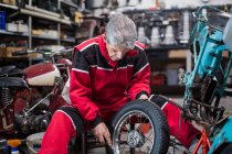 Побочный обзор серьезного квалифицированного пожилого механика мужского пола в ремонте старомодного сломанного мотоцикла и фиксирующего колеса в мастерской — стоковое фото