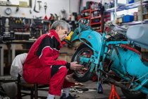 Vista lateral do mecânico idoso qualificado sério do sexo masculino no workwear que conserta a motocicleta quebrada antiquada e a roda de fixação na oficina — Fotografia de Stock