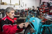 Side view of elderly gray haired male mechanic with screwdriver removing dirt and rust from broken retro motorcycle while working in garage — Stock Photo