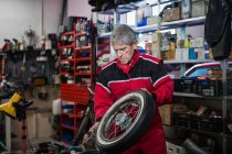 Reparador senior serio que fija la rueda de la motocicleta vieja mientras que trabaja en taller de servicio profesional - foto de stock