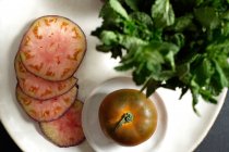 Vista superior de tomates negros frescos enteros y en rodajas sobre la mesa y la menta verde durante la preparación de comidas saludables - foto de stock