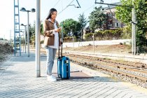 Зміст етнічних мандрівників, що стоять з валізою на платформі під час перегляду залізниць на смартфоні — стокове фото