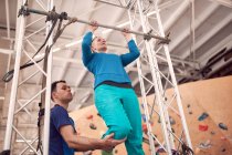 Снизу женщины делают подтягивания на горизонтальной полосе с помощью инструктора во время тренировки в современном альпинистском центре — стоковое фото