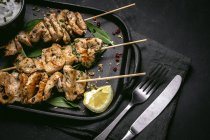 De dessus de viande fraîchement cuite appétissante sur brochettes servies sur plateau sur table noire avec bol de sauce — Photo de stock