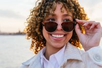 Mulher afro-americana alegre em óculos de sol elegantes em pé na praia e desfrutando de liberdade ao pôr do sol olhando para longe — Fotografia de Stock