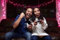 Casal étnico excitado em roupas casuais com almofadas de alegria jogando videogame juntos enquanto sentados no sofá de couro em casa — Fotografia de Stock