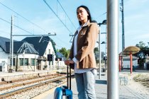 De baixo vista lateral do viajante feminino asiático com mala de pé na plataforma da estação ferroviária enquanto espera o trem — Fotografia de Stock