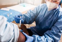 Medico donna ritagliato in uniforme protettiva e guanti in lattice che vaccina il paziente afroamericano maschio irriconoscibile in clinica durante l'epidemia di coronavirus — Foto stock