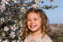 Criança sorrindo adorável no vestido que está perto da árvore florescente com flores no parque da mola e olhando afastado — Fotografia de Stock