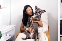 Conteúdo feminino étnico abraçando American Staffordshire Terrier com a língua para fora enquanto sentado em poltrona no quarto da casa — Fotografia de Stock