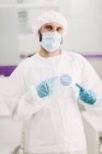 Positiver männlicher Arzt mit Latexhandschuhen und medizinischer Schutzmaske zeigt auf covid-19 geimpften Botschaftsaufkleber auf weißer Uniform, der in einer modernen Arztpraxis steht und in die Kamera blickt — Stockfoto