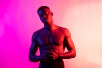 Serious young African American male athlete with naked torso looking at camera with hands folded on pink background in neon studio — Stock Photo